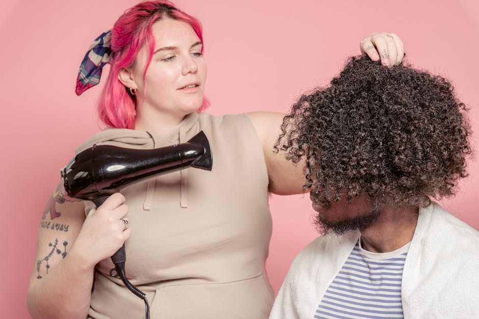 Female hairdresser with dyed hair drying hair of African American male model with Afro hairstyle against pink background
